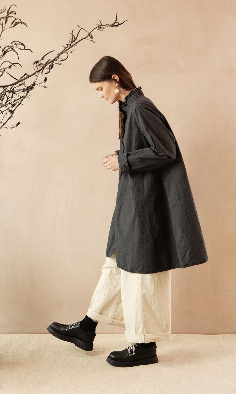 Plümo- Woman in a Cockle Shirt, wide cream pants, and black shoes walks indoors beside a bare branch on a neutral backdrop.