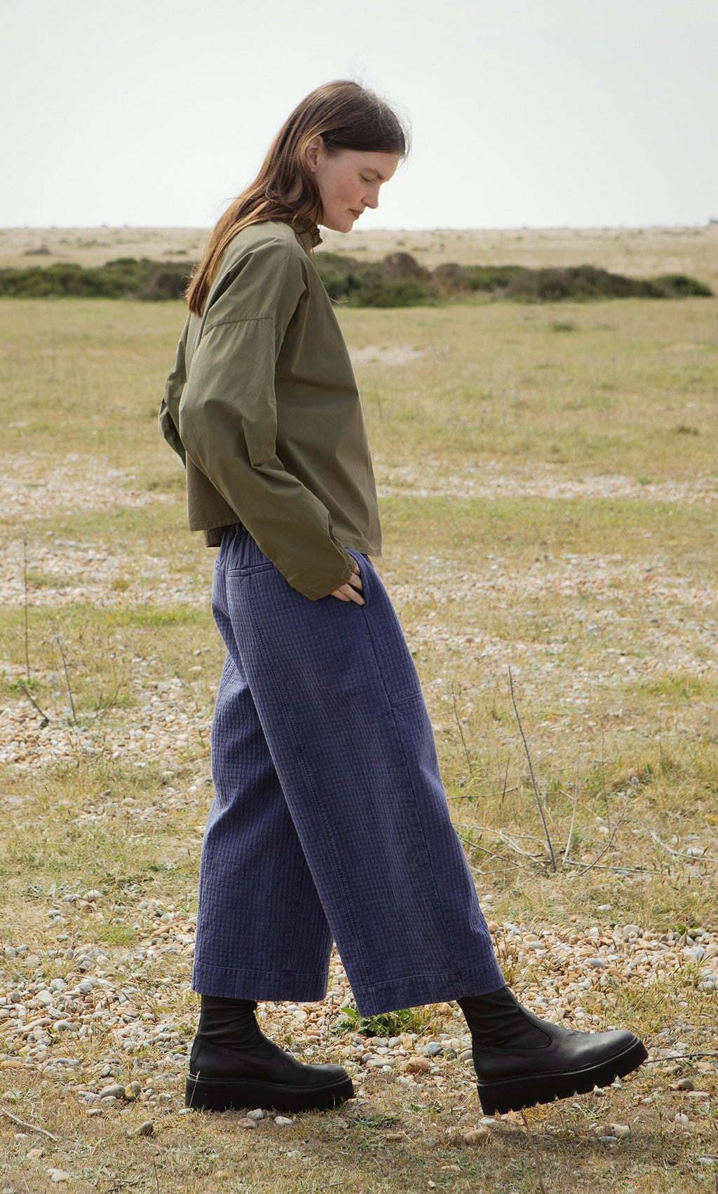 Plümo- Woman outdoors in an olive jacket, Calow Pants in checked fabric with curved legs, and black boots. Made in London.