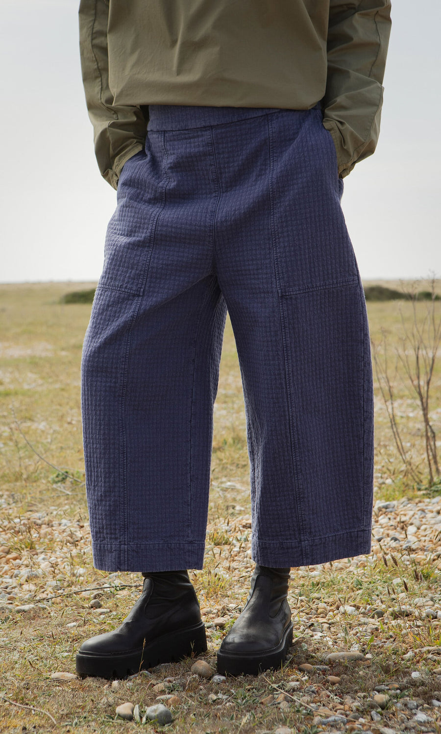 Plümo- Person poses outdoors in Calow Pants, black boots, and an olive jacket; face is out of frame.