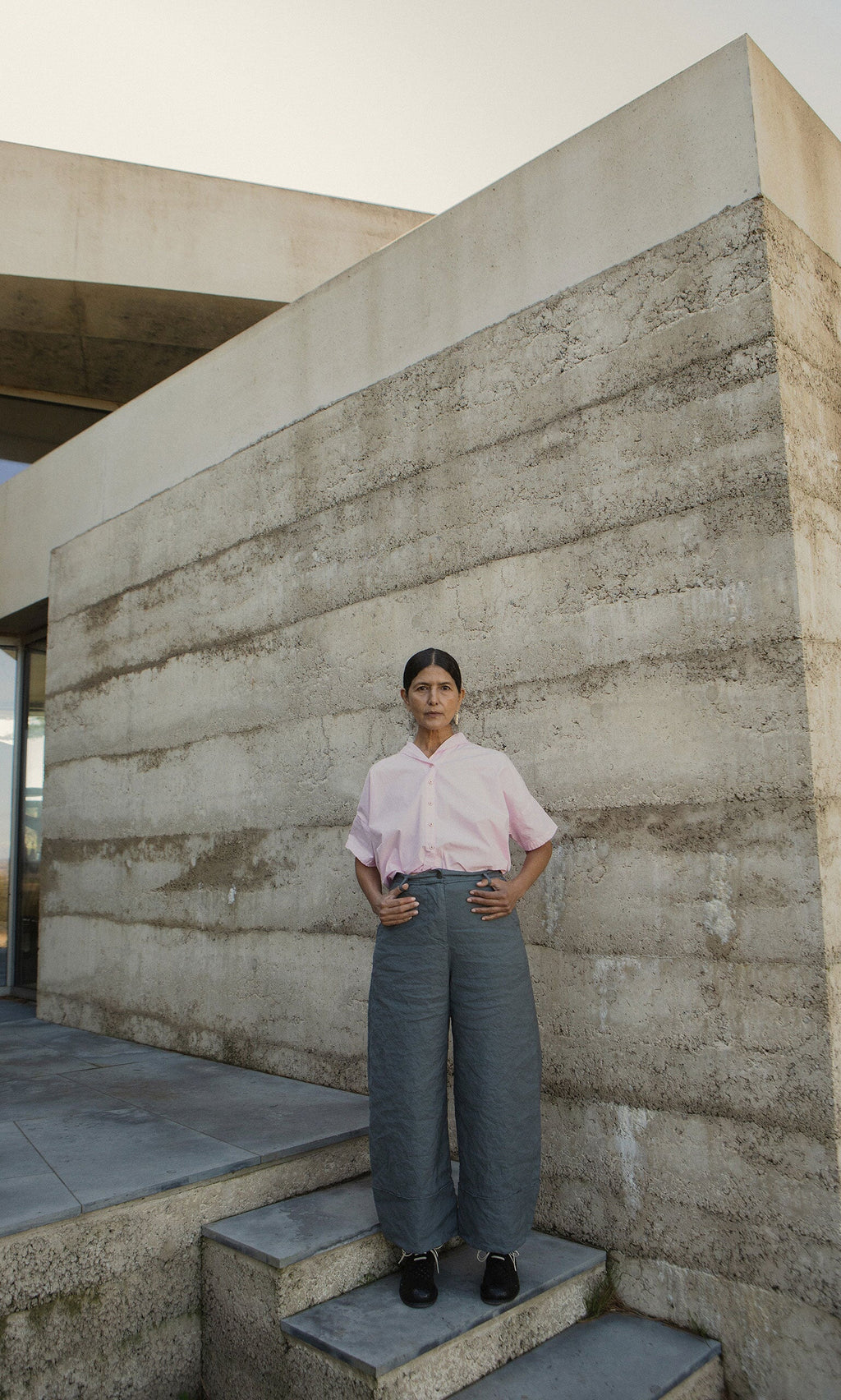 Plümo- A person stands on steps before a modern building, wearing a pink shirt and the Cora Grey Pants.
