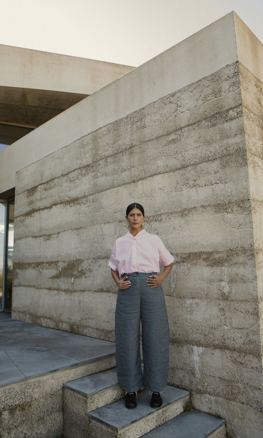 Plümo- A person stands on steps before a modern building, wearing a pink shirt and the Cora Grey Pants.