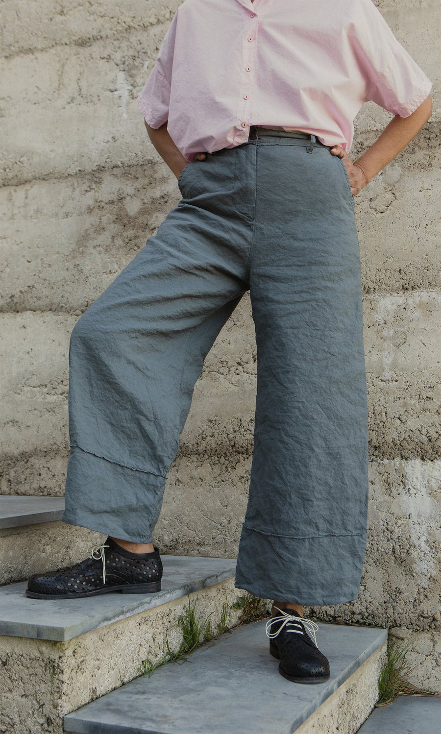 Plümo- Person wearing a pink shirt, Cora Grey Pants, and black shoes stands on concrete steps against a stone wall.