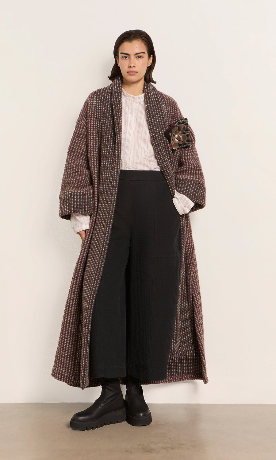 Plümo  A person in a Cordova Coat, white shirt, and black pants stands against a plain wall.