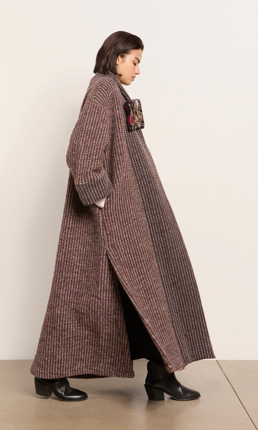 Plümo  Person in a Cordova Coat and black boots, with a scarf, standing sideways against a plain indoor wall.