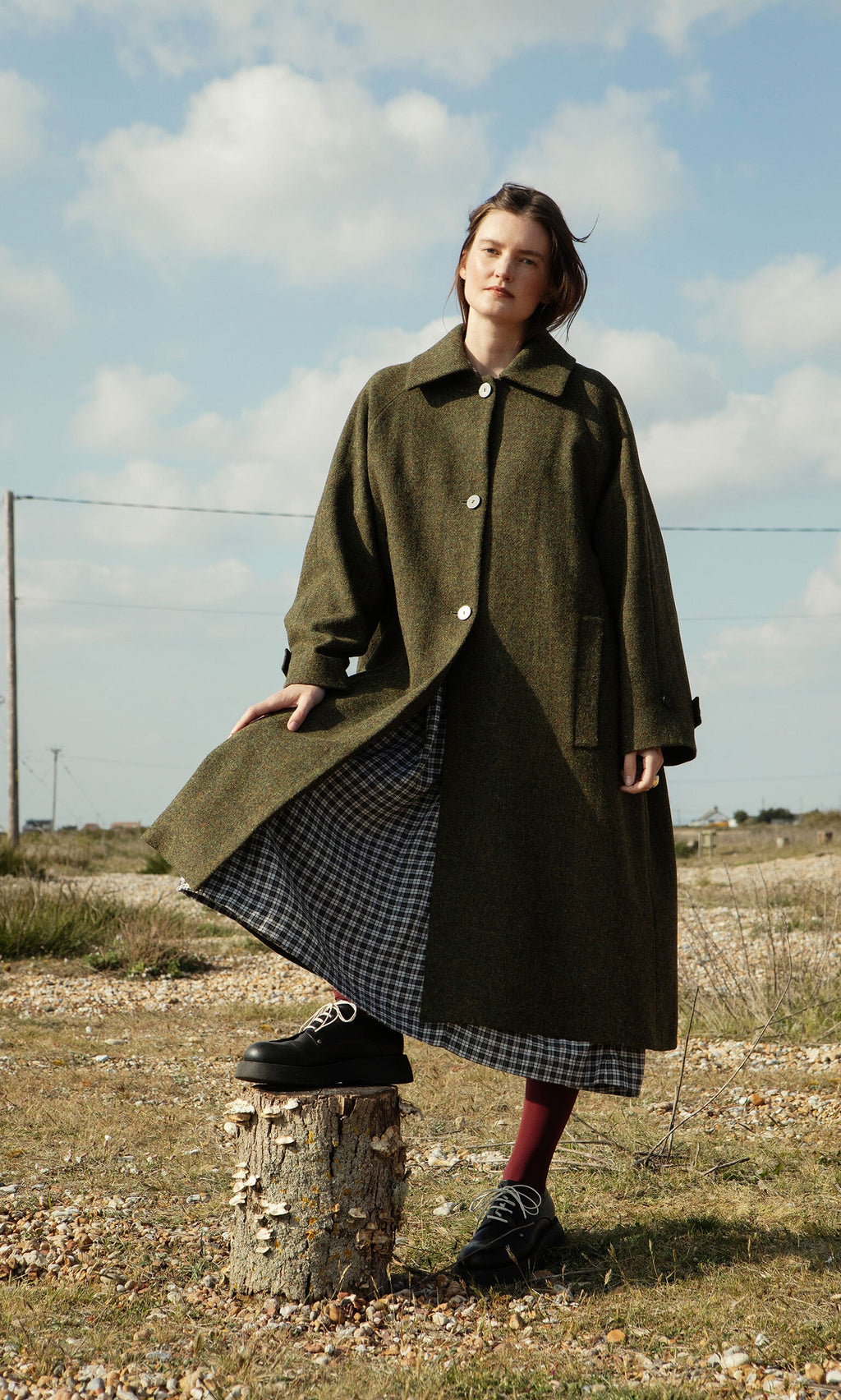 Plümo- Woman wears the Cormack Coat - Harris Tweed and a checkered dress outdoors, one foot on a tree stump under cloudy skies.