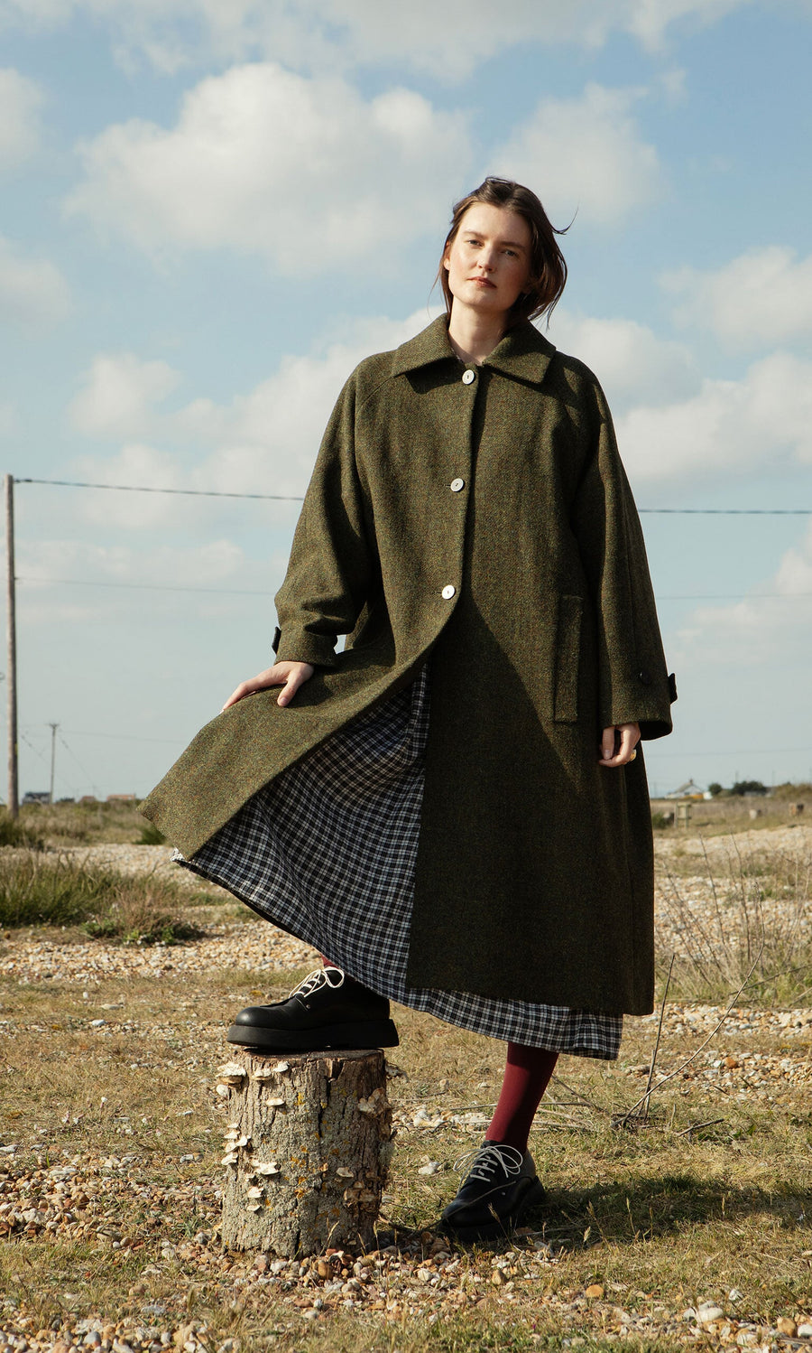 Plümo- Woman wears the Cormack Coat - Harris Tweed and a checkered dress outdoors, one foot on a tree stump under cloudy skies.