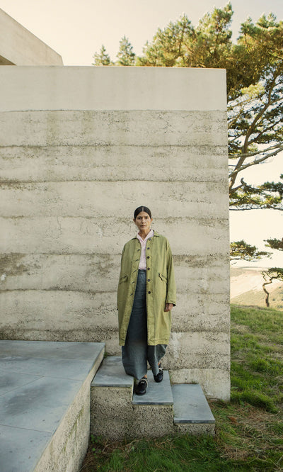 Plümo- Person in a hand-dyed Cragside Coat stands on stone steps by a modern building, with grass and trees behind.