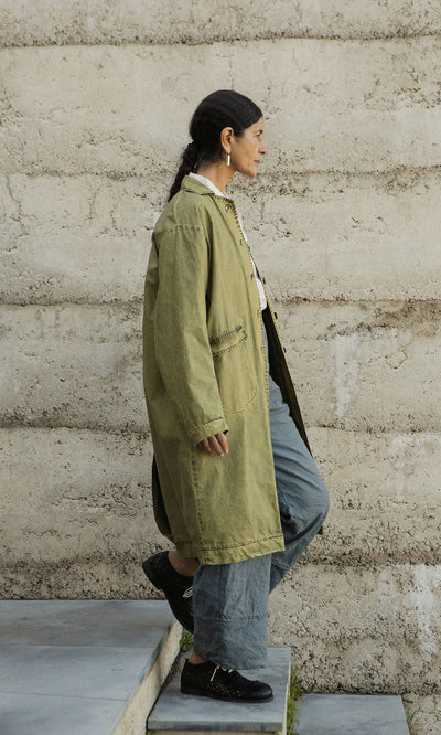 Plümo- Wearing a hand-dyed Cragside Coat and gray pants, a person walks up concrete steps next to a textured beige wall.
