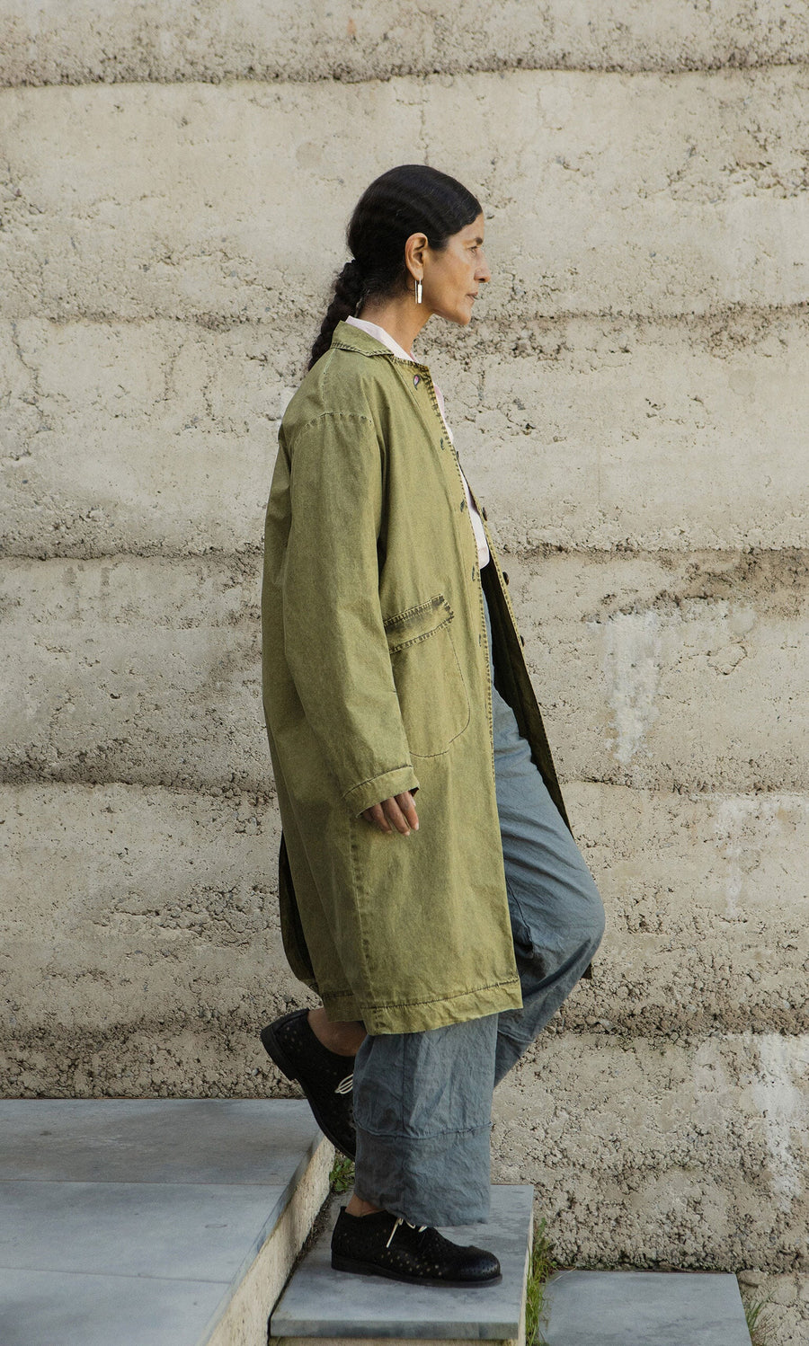 Plümo- Wearing a hand-dyed Cragside Coat and gray pants, a person walks up concrete steps next to a textured beige wall.