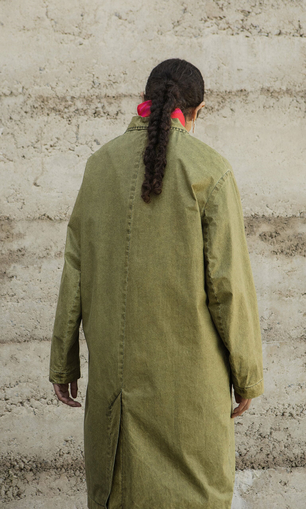 Plümo- Person with long braided hair in an olive green Cragside Coat stands facing a textured beige wall.