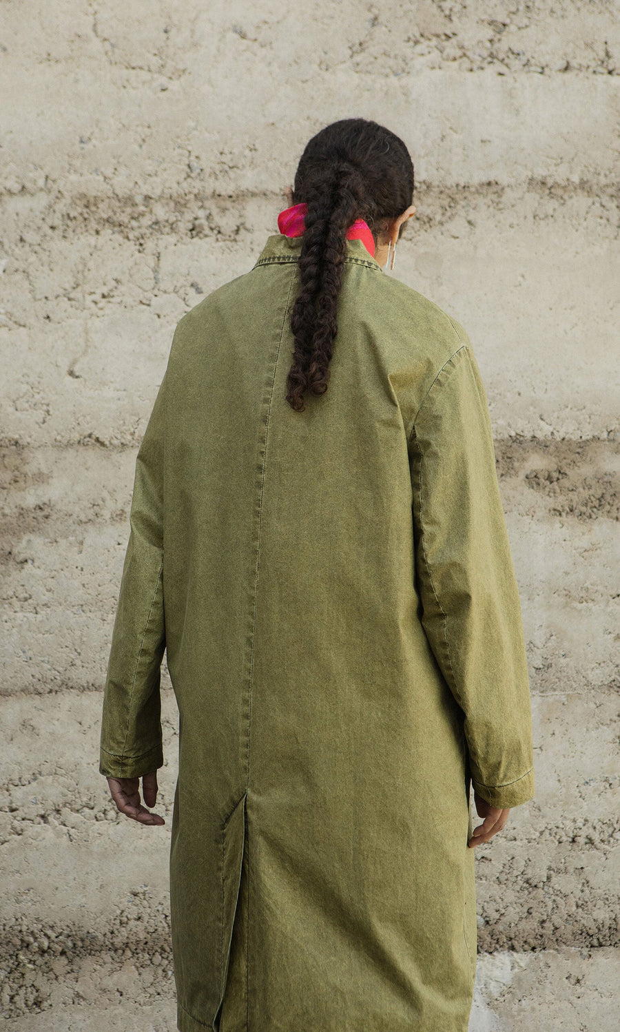 Plümo- Person with long braided hair in an olive green Cragside Coat stands facing a textured beige wall.