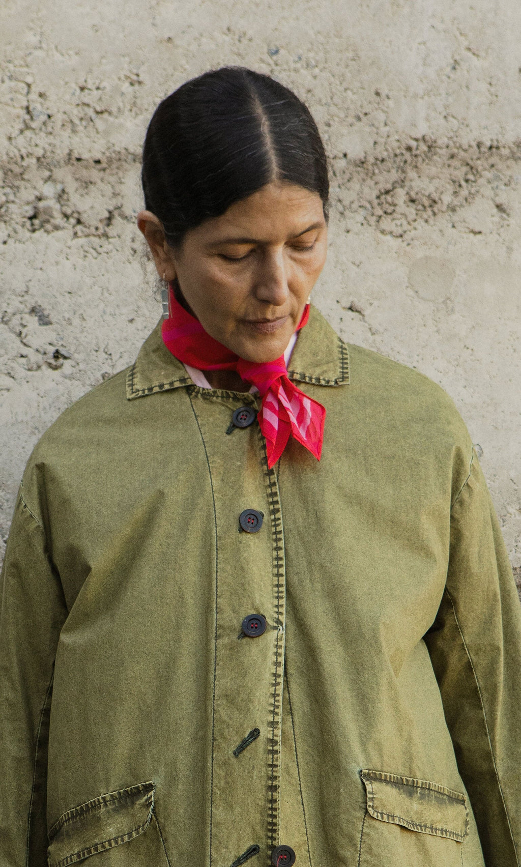 Plümo- Person with dark hair in a green jacket and Poinsettia Necktie looks downward before a textured concrete wall.