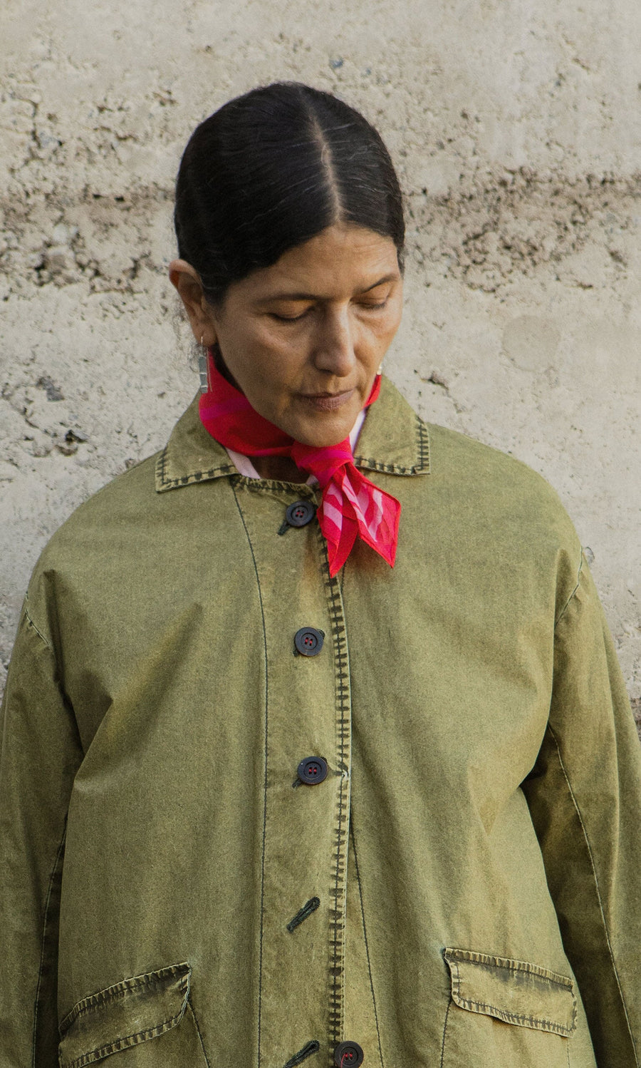 Plümo- Person with dark hair in a green jacket and Poinsettia Necktie looks downward before a textured concrete wall.