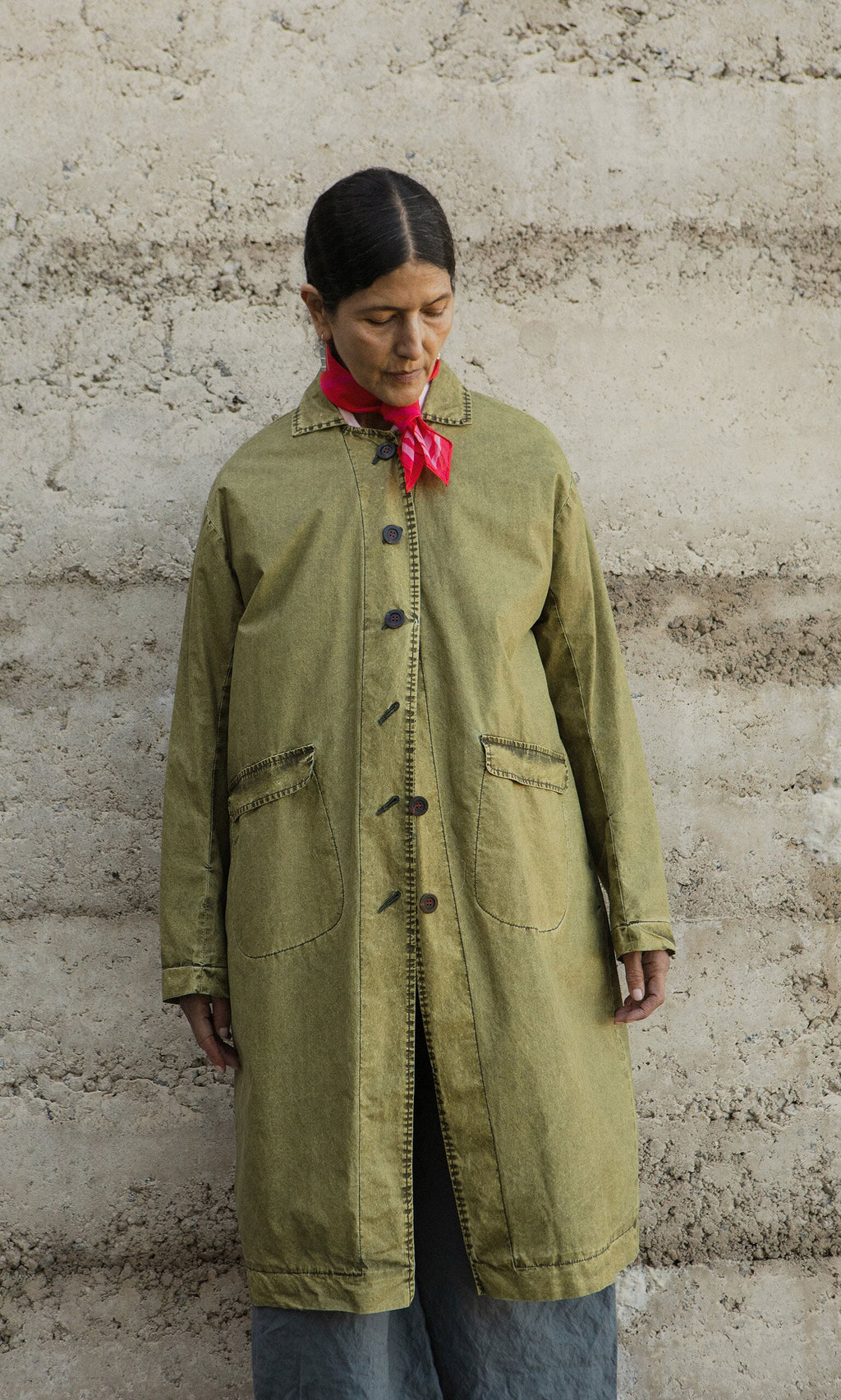 Plümo- A person in a hand-dyed Cragside Coat and red scarf stands against a textured beige wall, looking down.