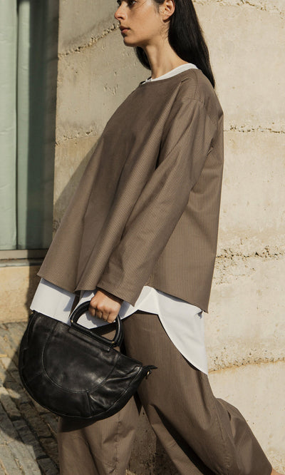 Plümo- A woman in taupe oversized clothing carries the Dark Moon Tote and walks past a stone wall.
