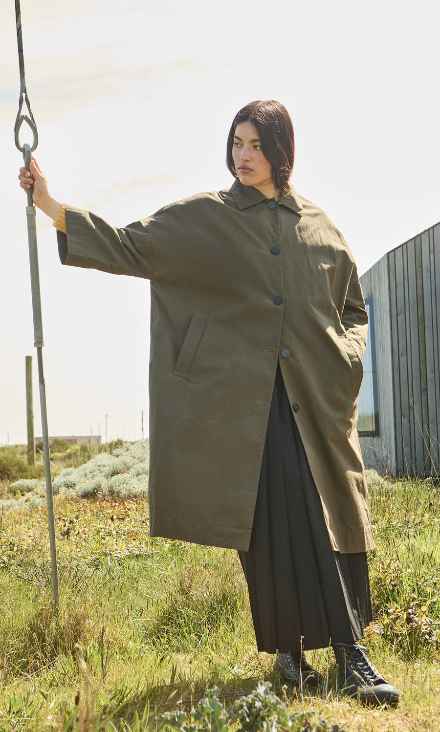 Plümo- Person in a Doherty Coat - Olive stands on grass, holding a pole, with a modern curved building behind them.