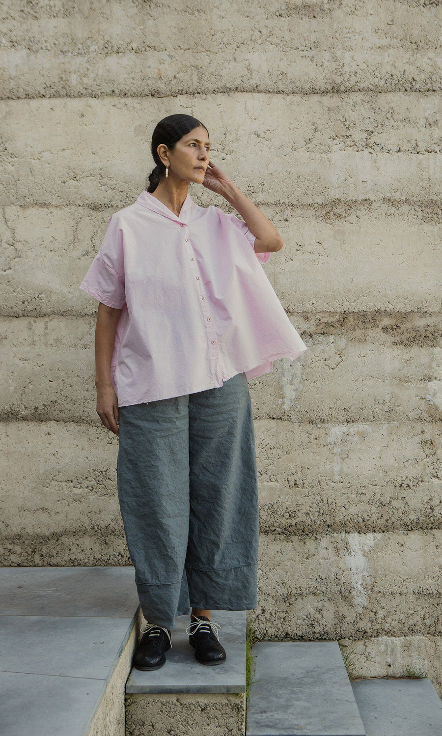 Plümo- Woman in the Early Pink Blouse by Hannoh Wessel stands on concrete steps by a beige wall, looking thoughtful.