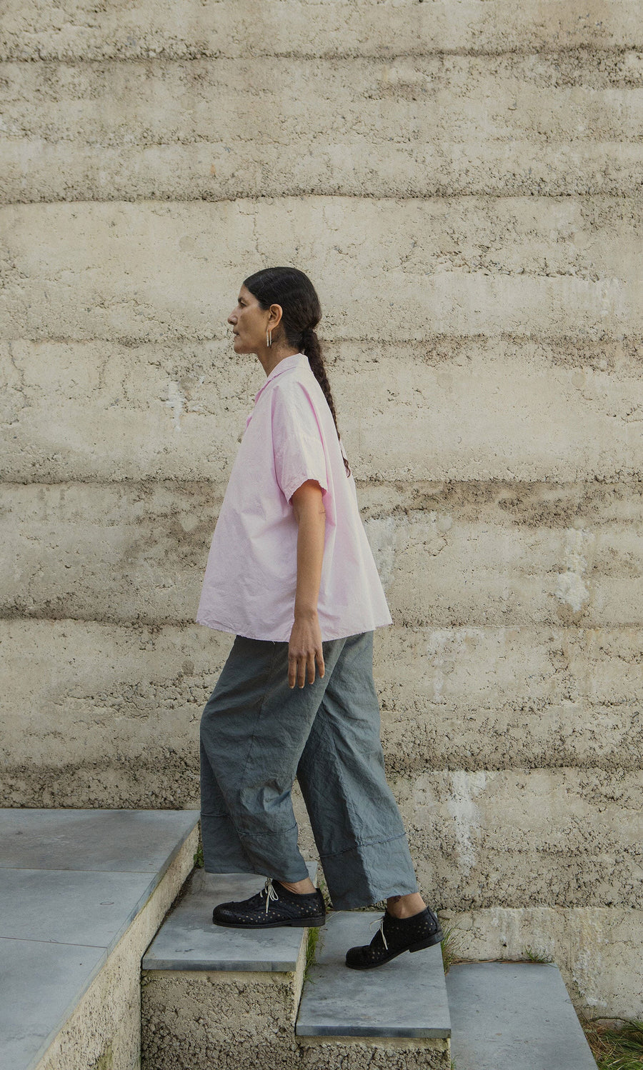 Plümo- Someone in a pink shirt and Cora Grey Pants walks up gray steps next to a textured beige wall.