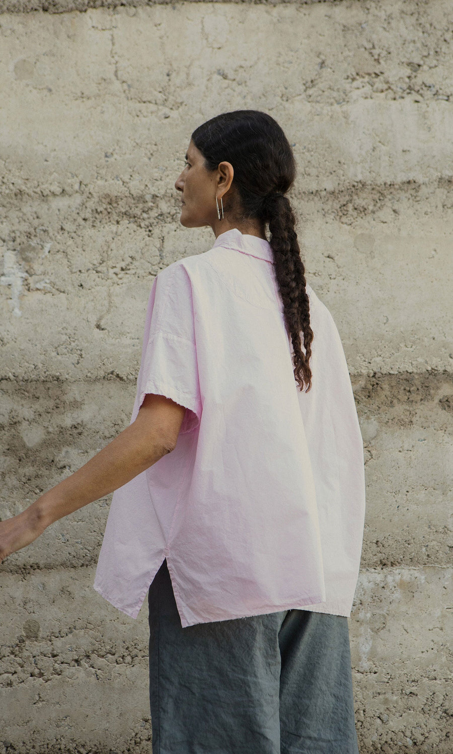 Plümo- Person with a long braid wears the Early Pink Blouse, standing by a textured beige wall.