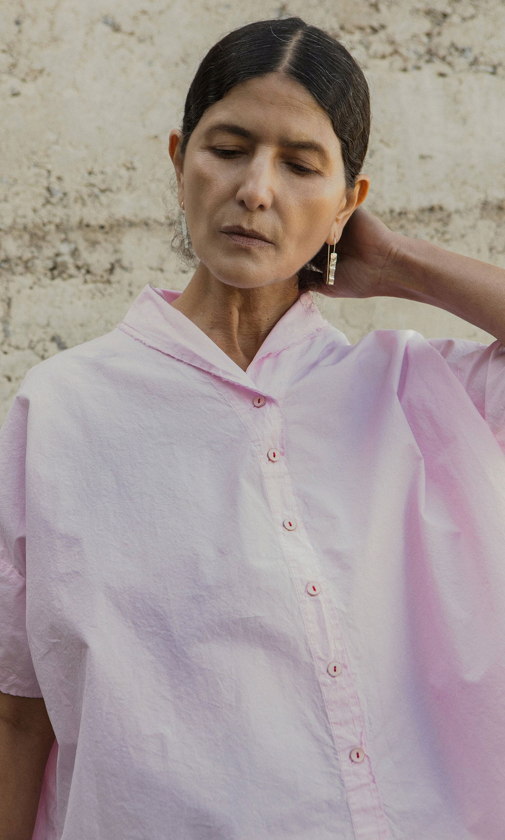 Plümo- Woman in an Early Pink Blouse by Hannoh Wessel stands against a light wall, looking down and touching her neck.