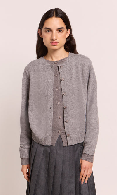 Plümo  Someone wears a Grey Melange Cardigan and pleated skirt, standing before a plain, light-colored backdrop.