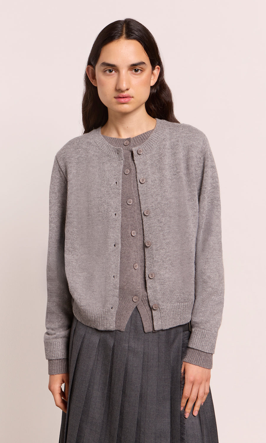 Plümo  Someone wears a Grey Melange Cardigan and pleated skirt, standing before a plain, light-colored backdrop.
