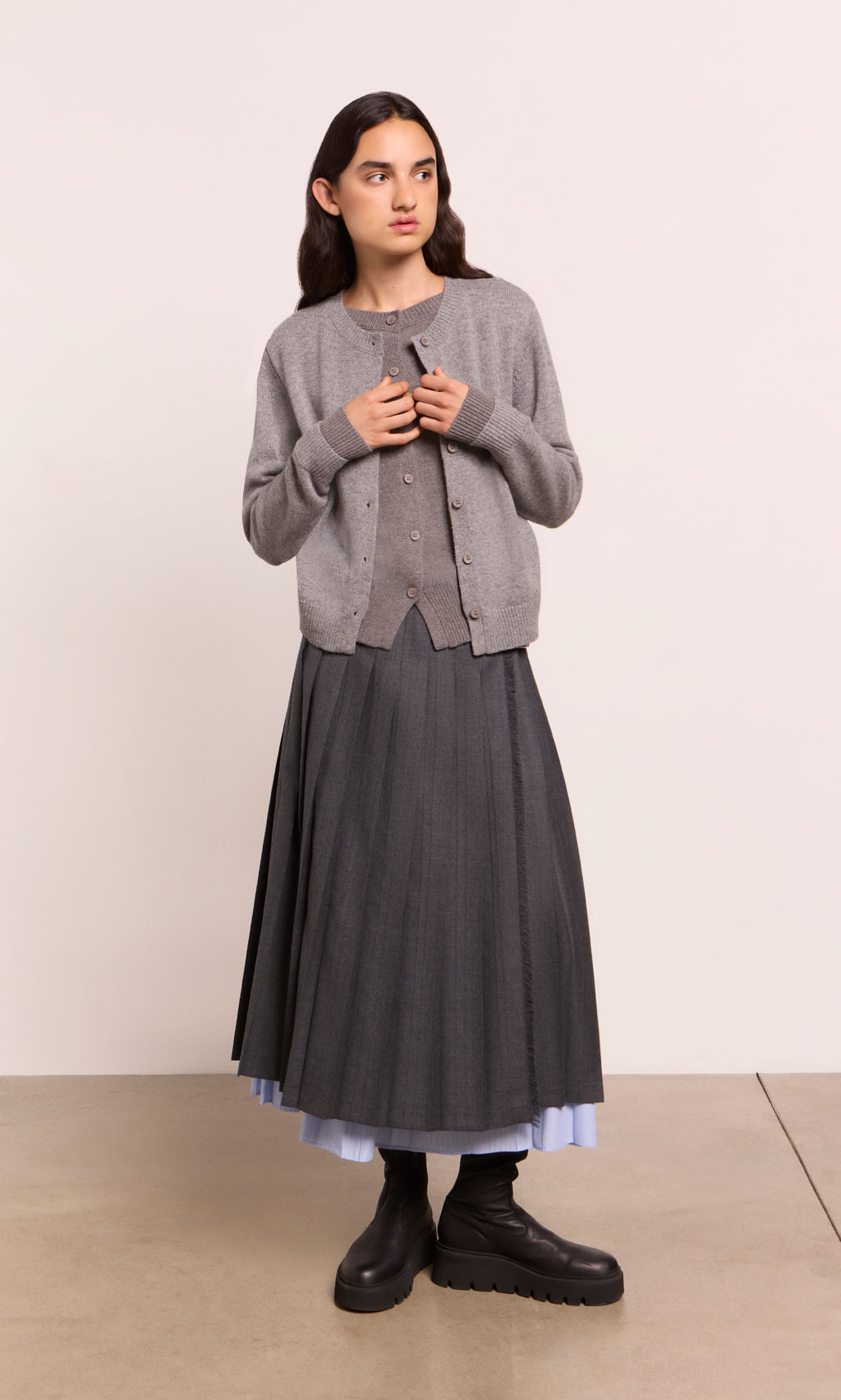 Plümo  Person in a Grey Melange Cardigan, long pleated skirt, black boots, on a light-colored floor.
