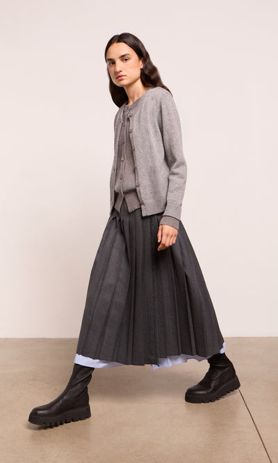Plümo  Person in a gray cardigan, Boyd Kilt, and black boots walks indoors against a plain background.