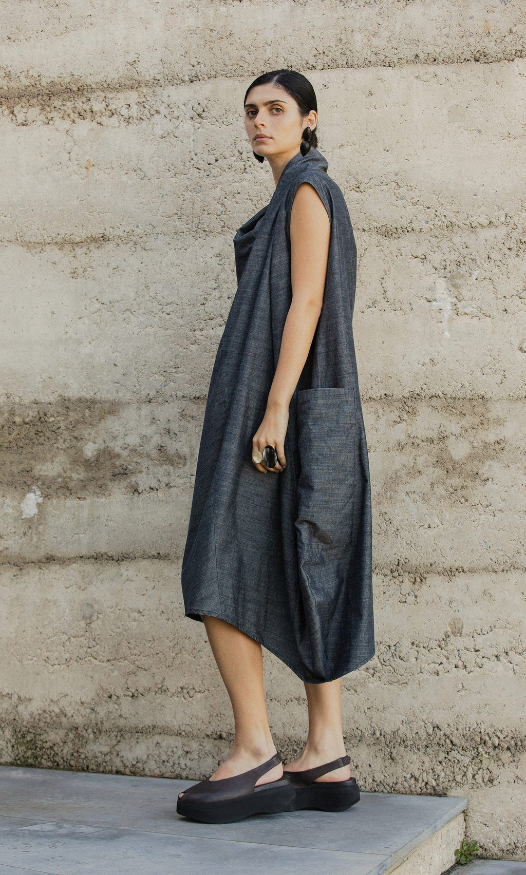 Plümo- Woman wearing the Emi Dress with large pocket, standing sideways by a textured concrete wall.