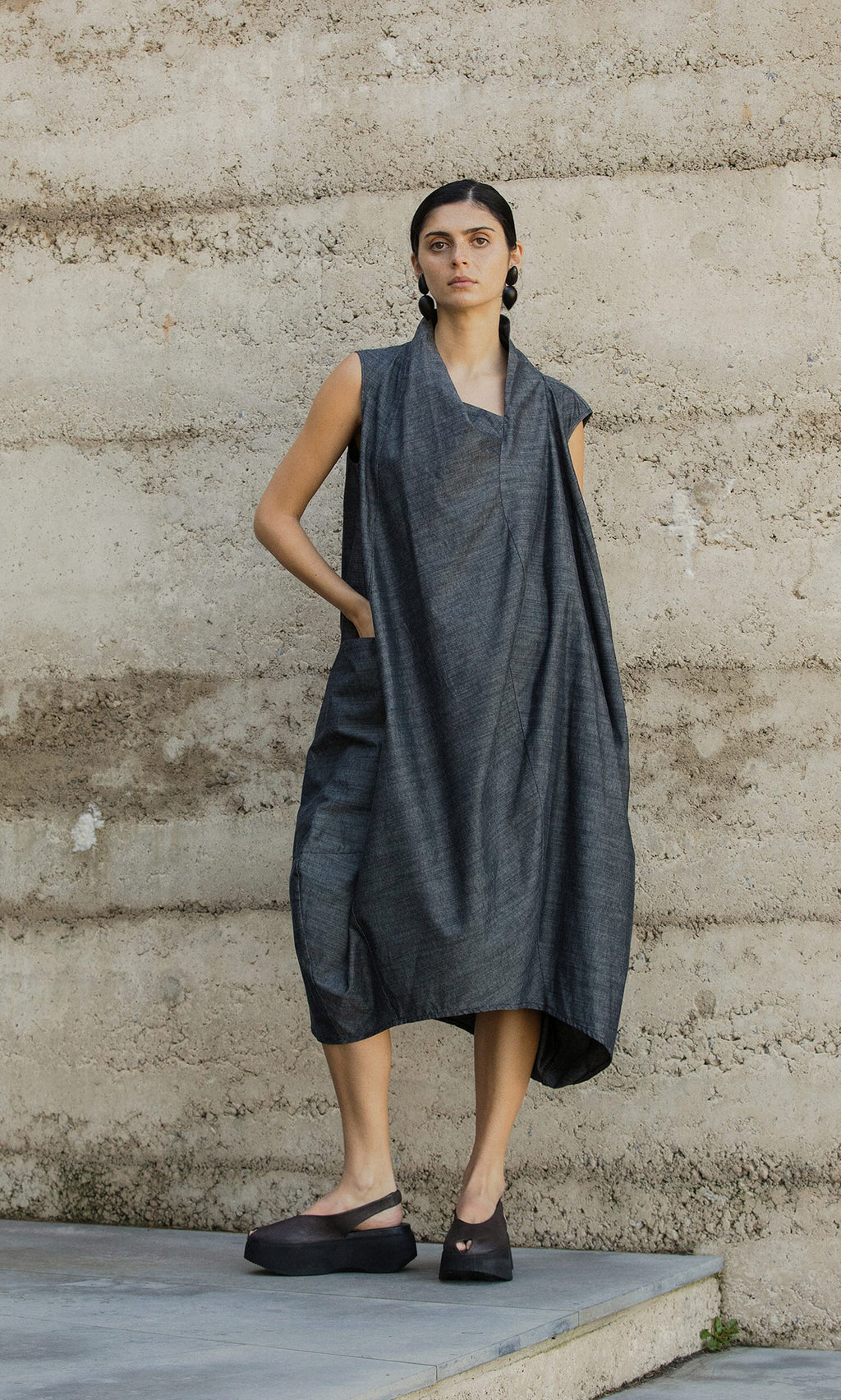 Plümo- A woman in the Emi Dress and black platform shoes stands against a textured concrete wall.