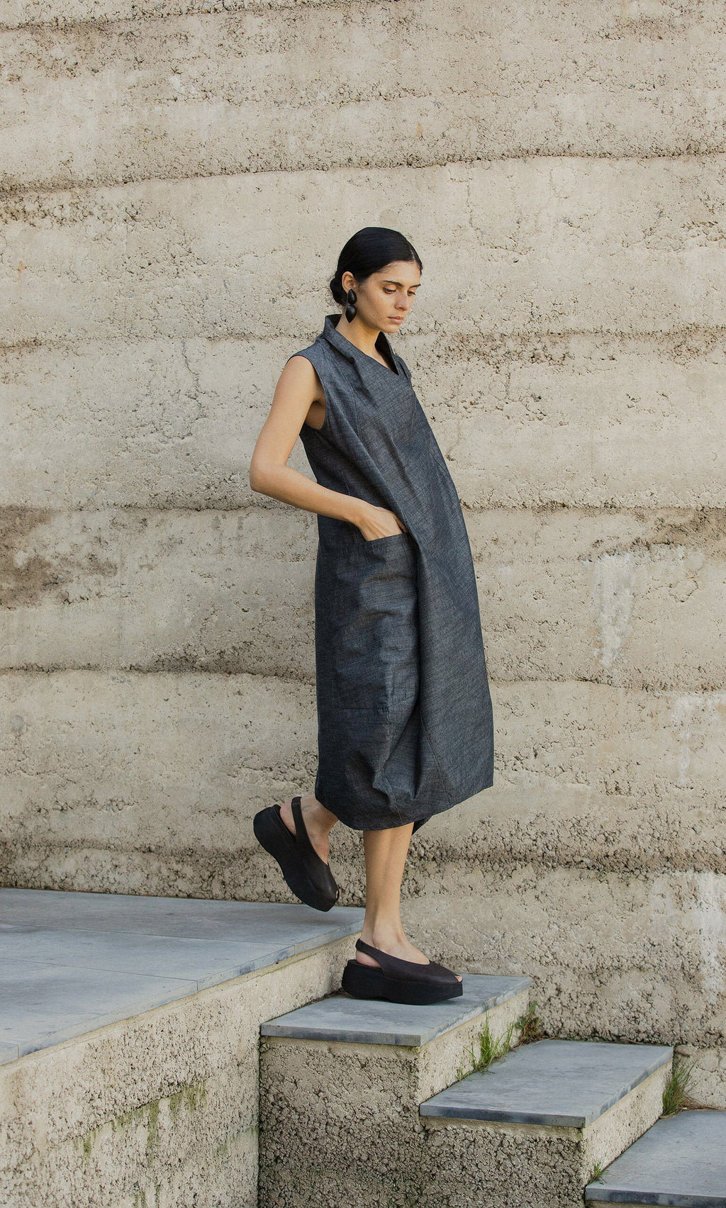 Plümo- Woman in the egg-shaped Emi Dress and black platform shoes stands on concrete stairs beside a textured beige wall.