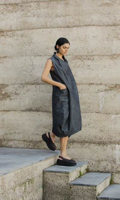 Plümo- Woman in the egg-shaped Emi Dress and black platform shoes stands on concrete stairs beside a textured beige wall.