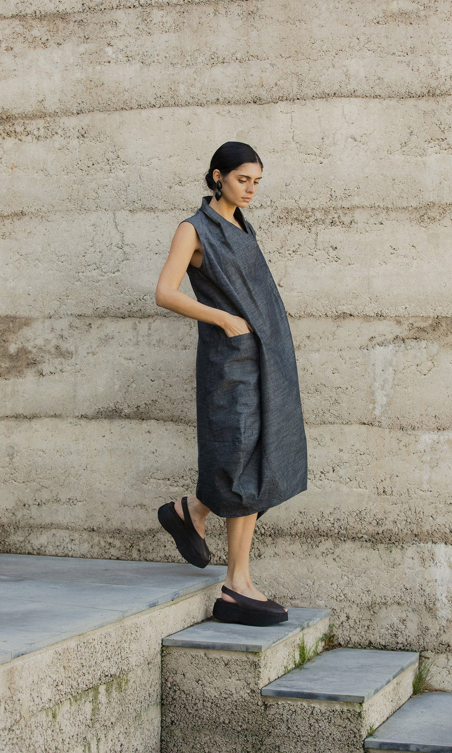 Plümo- Woman in the egg-shaped Emi Dress and black platform shoes stands on concrete stairs beside a textured beige wall.