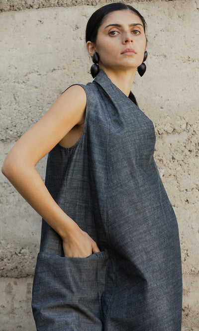 Plümo- Woman in the sleeveless gray Emi Dress with hand in pocket, standing against a textured concrete wall.