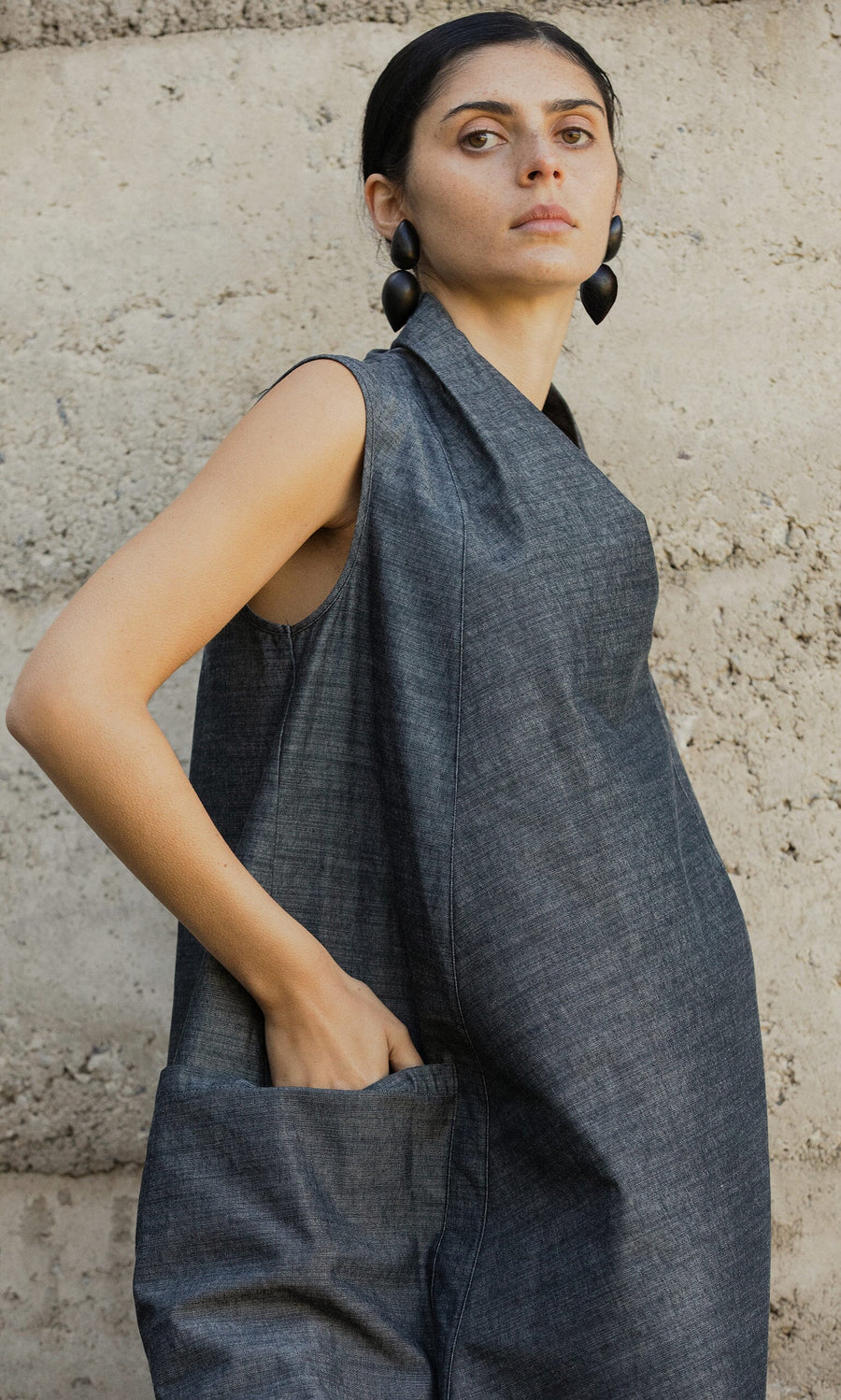 Plümo- Woman in the sleeveless gray Emi Dress with hand in pocket, standing against a textured concrete wall.