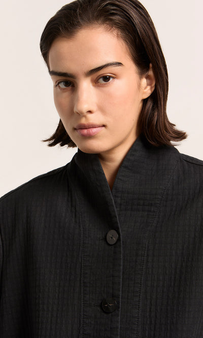 Plümo  Person with short dark hair wears the Envoy Jacket - Coal, looking at the camera with a neutral expression.