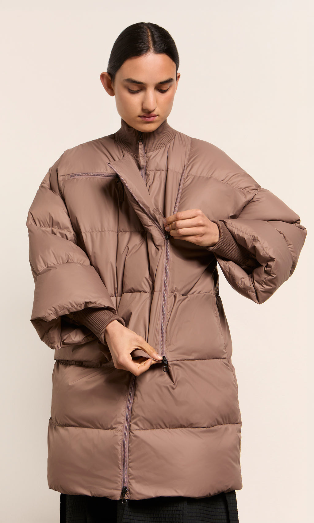 Plümo  Person zipping up the Firth Puffer Coat in fawn, looking down against a neutral background.