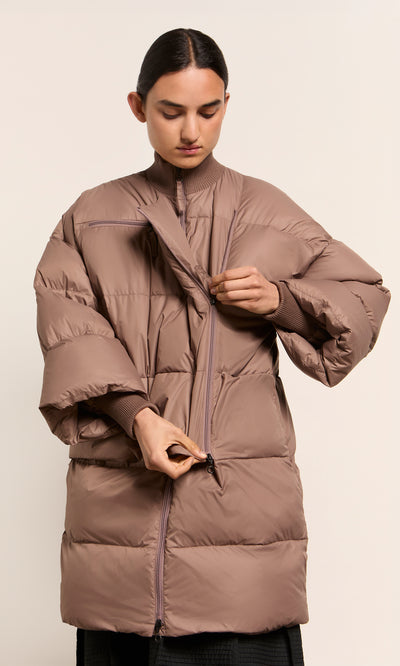Plümo  Person zipping up the Firth Puffer Coat in fawn, looking down against a neutral background.