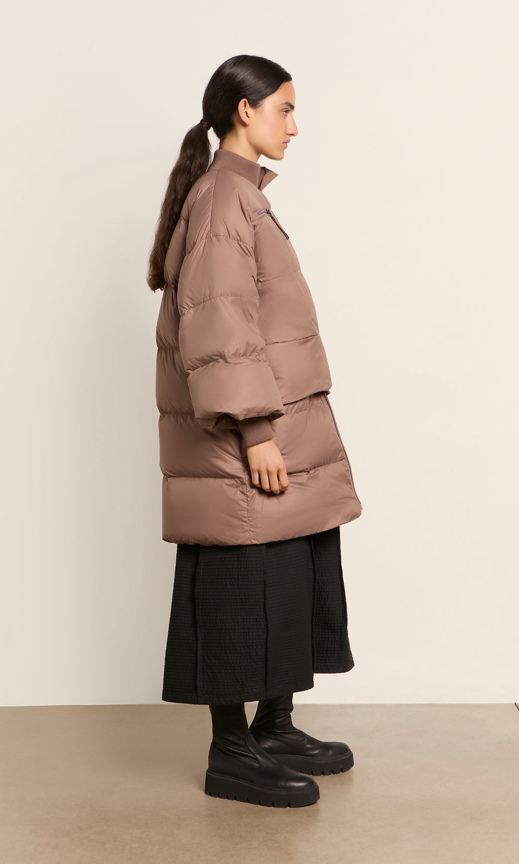 Plümo  Woman in a Firth Puffer Coat, black skirt, and boots stands sideways against a plain background.