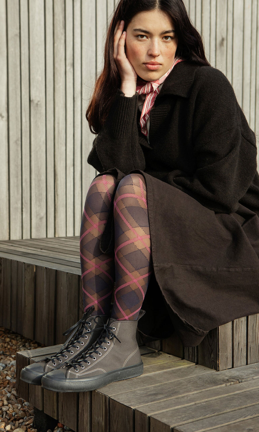 Plümo- Young woman in a black coat and patterned tights sits on steps, wearing High Top Trainers - Charcoal, looking at camera.