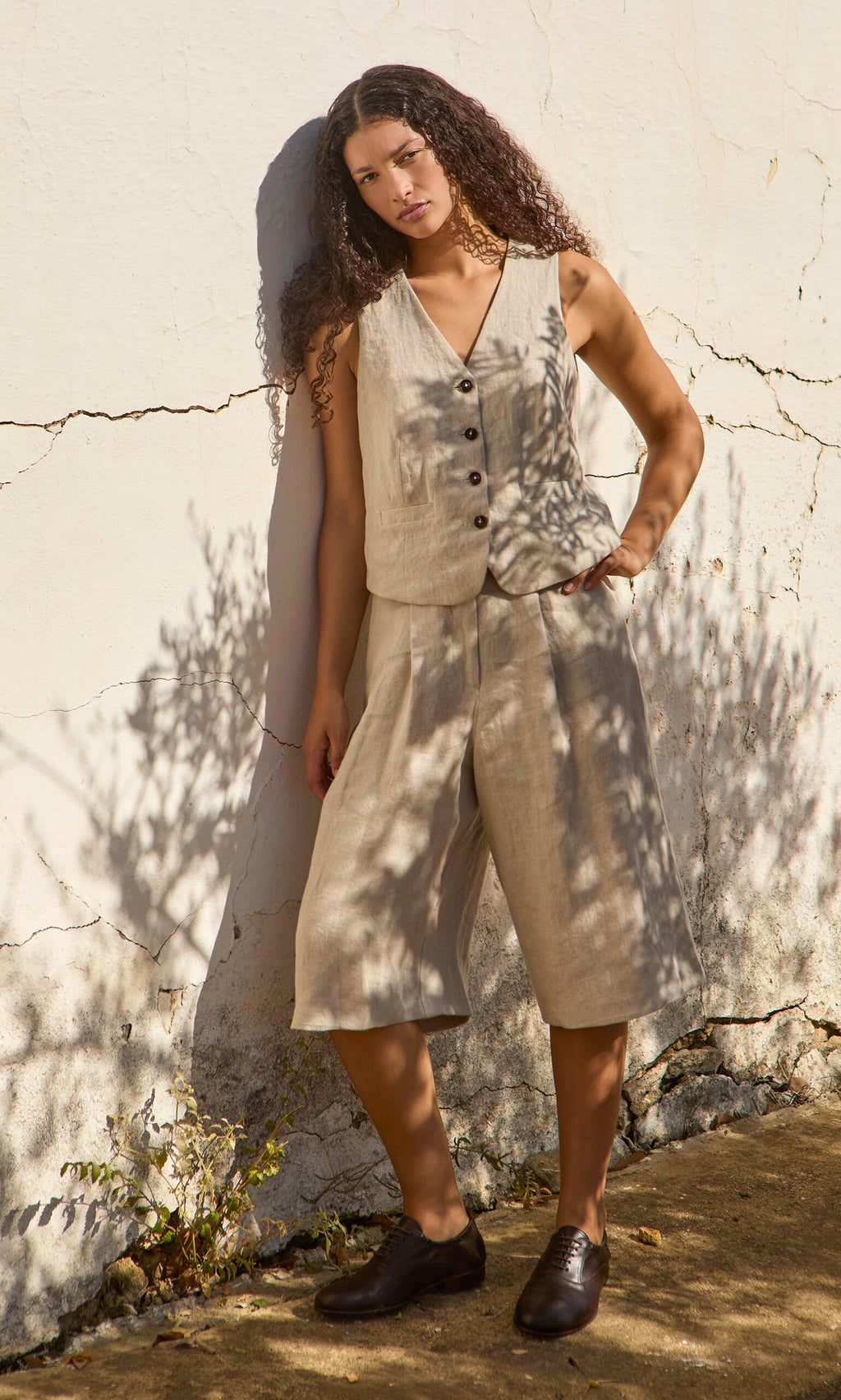 Plümo- Woman wearing Lucien Shorts - Natural stands by a sunlit wall, leaf shadows playing on her beige linen outfit.
