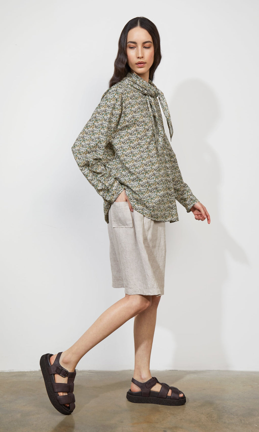 Plümo- A person in a Garden Blouse, beige shorts, and black sandals stands against a plain white wall.