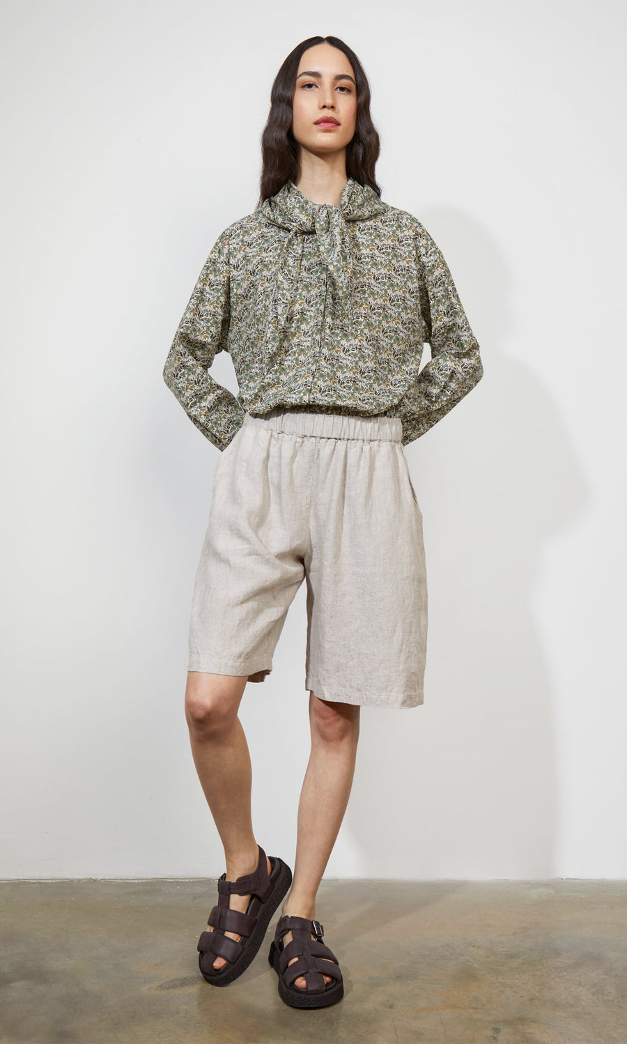 Plümo- Woman stands against a white wall wearing the Garden Blouse, beige shorts, and brown sandals with hands in pockets.