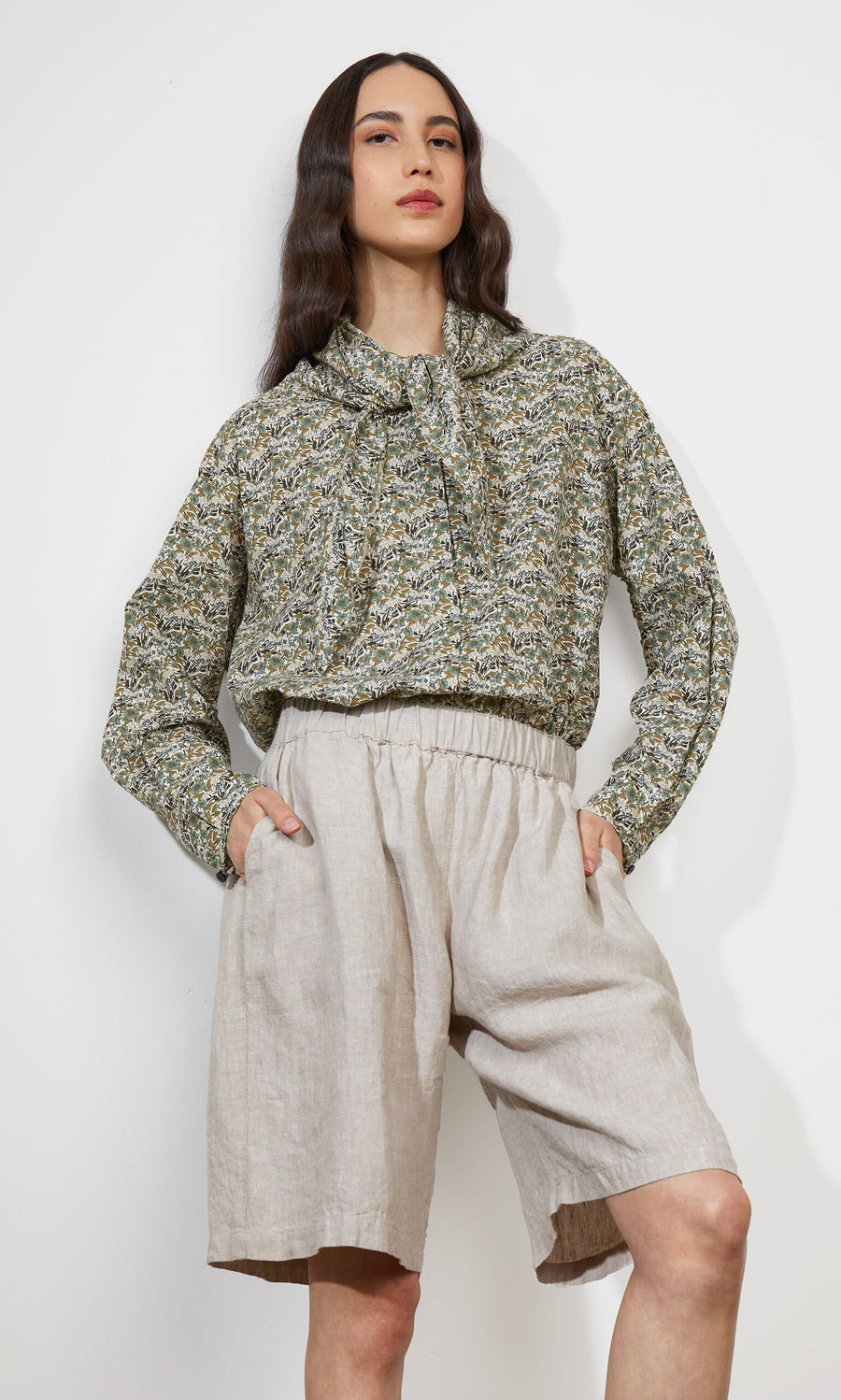 Plümo- Woman in Garden Blouse with neckerchief tie and light beige high-waisted shorts, hands in pockets.