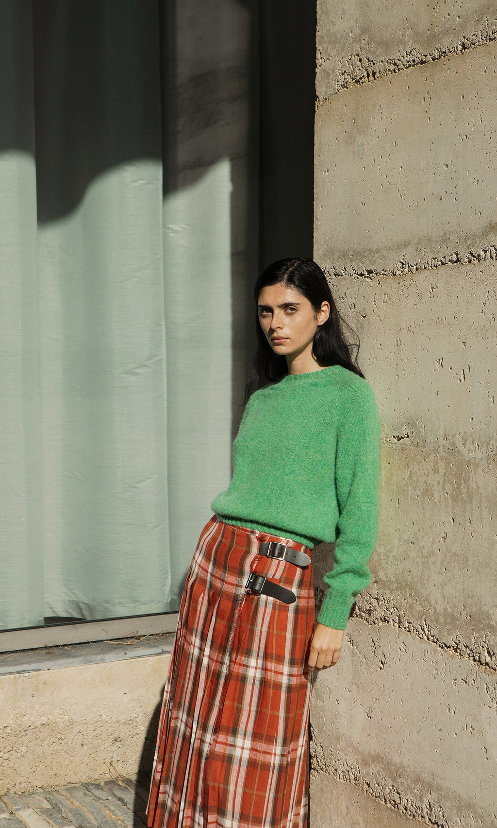 Plümo- Woman in a Wicklow Crewneck - Garden Leaf sweater with a red plaid skirt stands before pale green curtains.