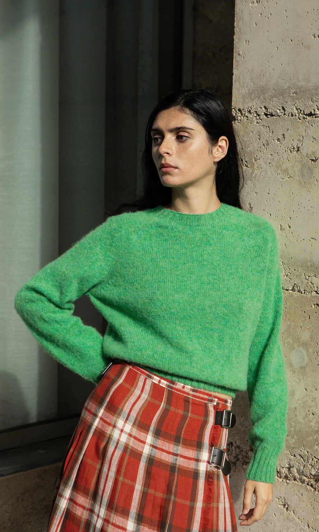 Plümo- Woman in a Wicklow Crewneck - Garden Leaf sweater and red plaid skirt stands by a textured wall, looking to the side.