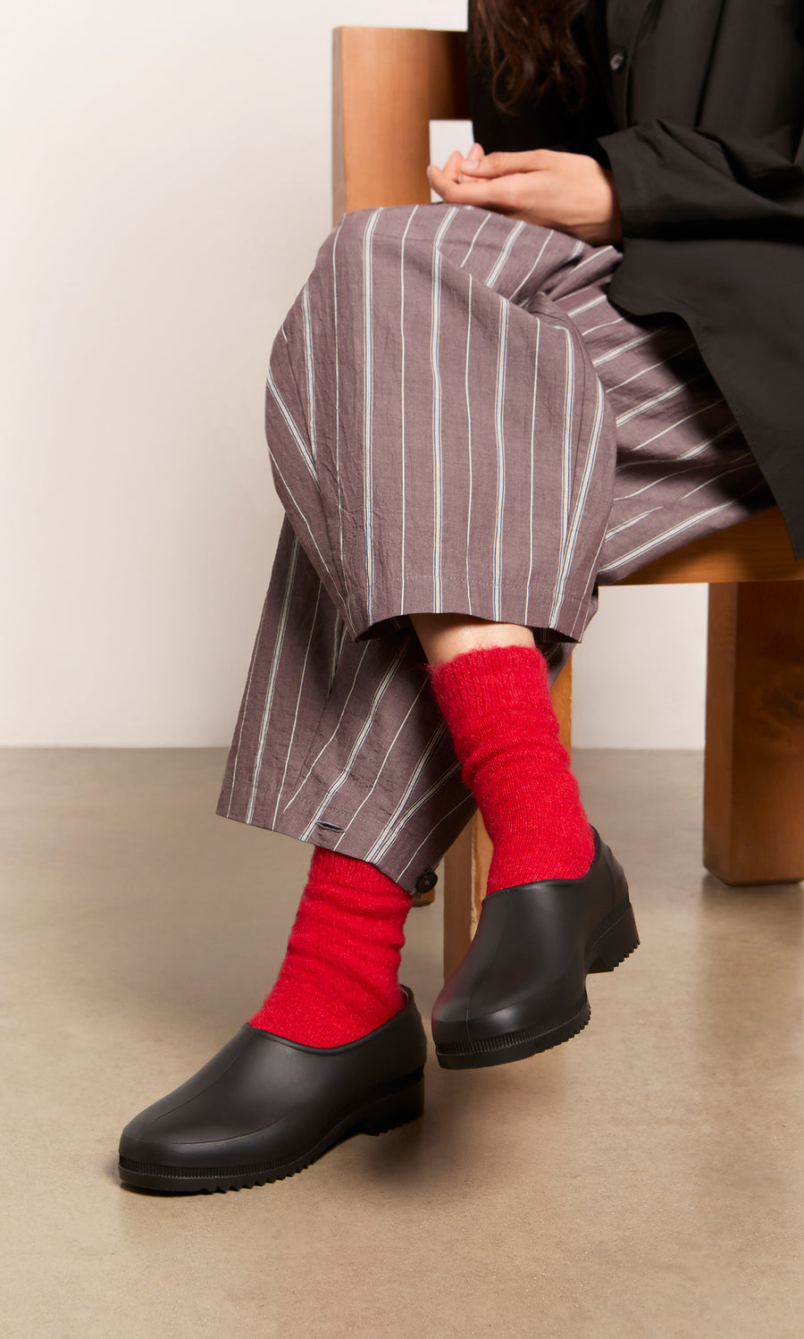 Plümo  Person seated in gray striped pants, red socks, and Gardenia Clogs - Black, hands resting on knee.