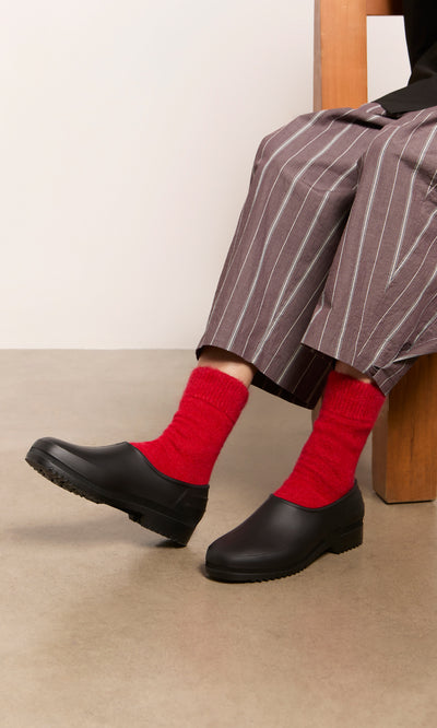 Plümo  Wearing Alpaca Socks - Postbox Red, black slip-ons, and pinstripe pants while seated on a wooden chair.