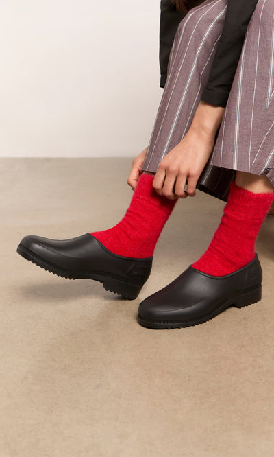 Plümo  Person in Alpaca Socks - Postbox Red and black shoes, hands near ankles, wearing striped pants.