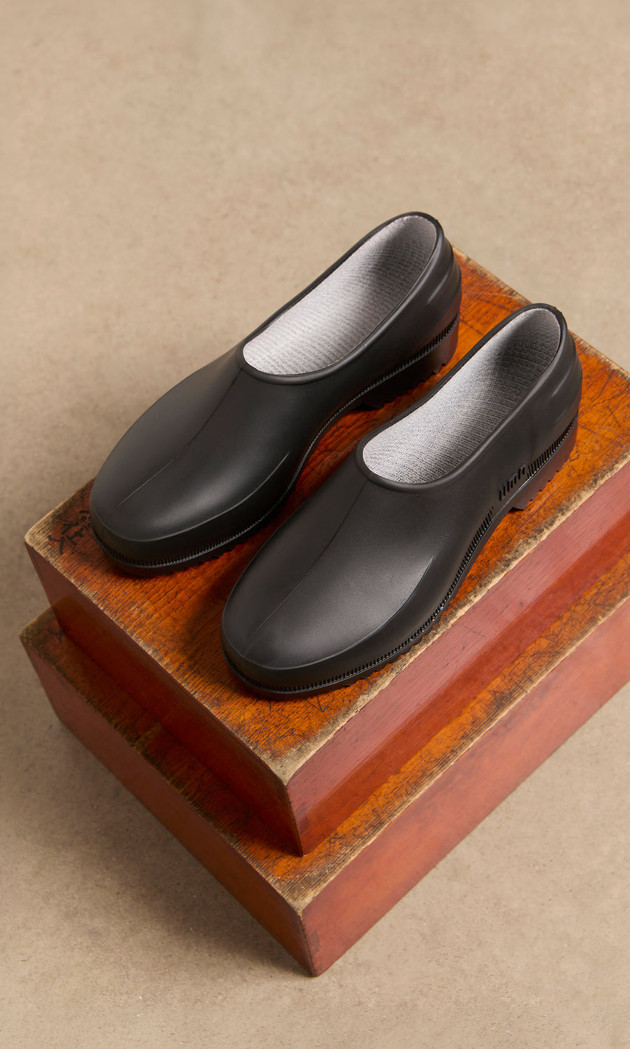 Plümo  Gardenia Clogs - Black with white lining on stacked wooden boxes, beige floor in the background.